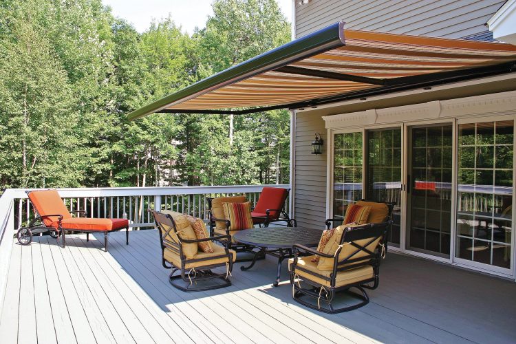 A spacious wooden deck with cushioned patio furniture, a table, and a red lounge chair. An extended striped retractable awning provides shade. Trees are visible in the background.
