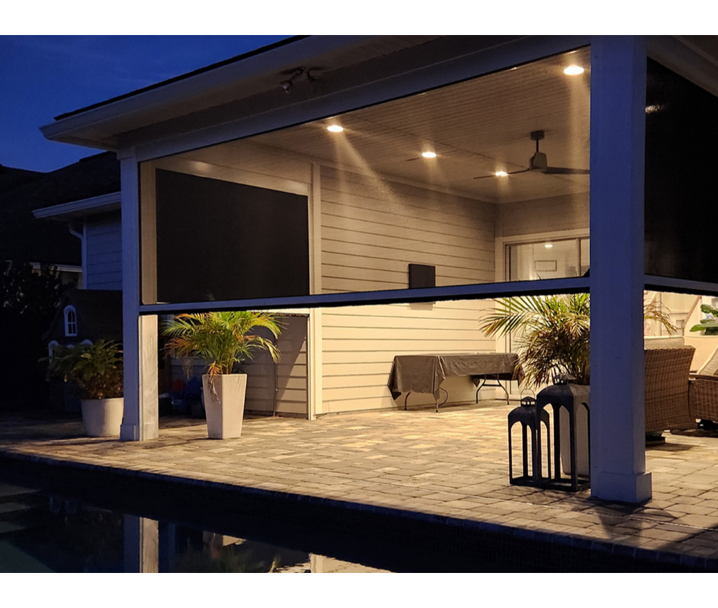 Beautiful motorized screen and a covered patio at dusk with potted plants, outdoor furniture, ceiling lights, and a view of a pool in the foreground. The area is illuminated by warm lighting, creating a cozy ambiance.