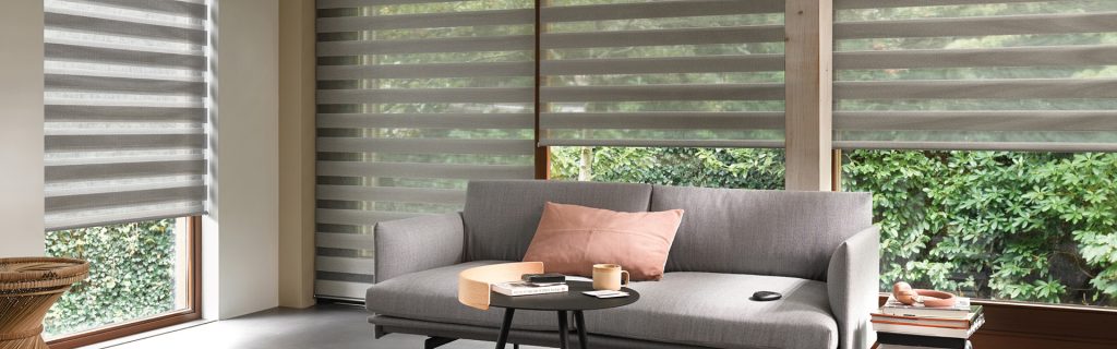 A modern living room with large windows covered by striped sheer blinds, a gray sofa with a pink pillow, a small round table with coffee, books, and remote, and greenery visible outside.