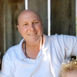 Chris Irminger in a white collared shirt relaxes beneath motorized awnings in a shaded outdoor area in Central Florida, with a blurred background. A person with curly blonde hair is partially visible in the lower right corner.