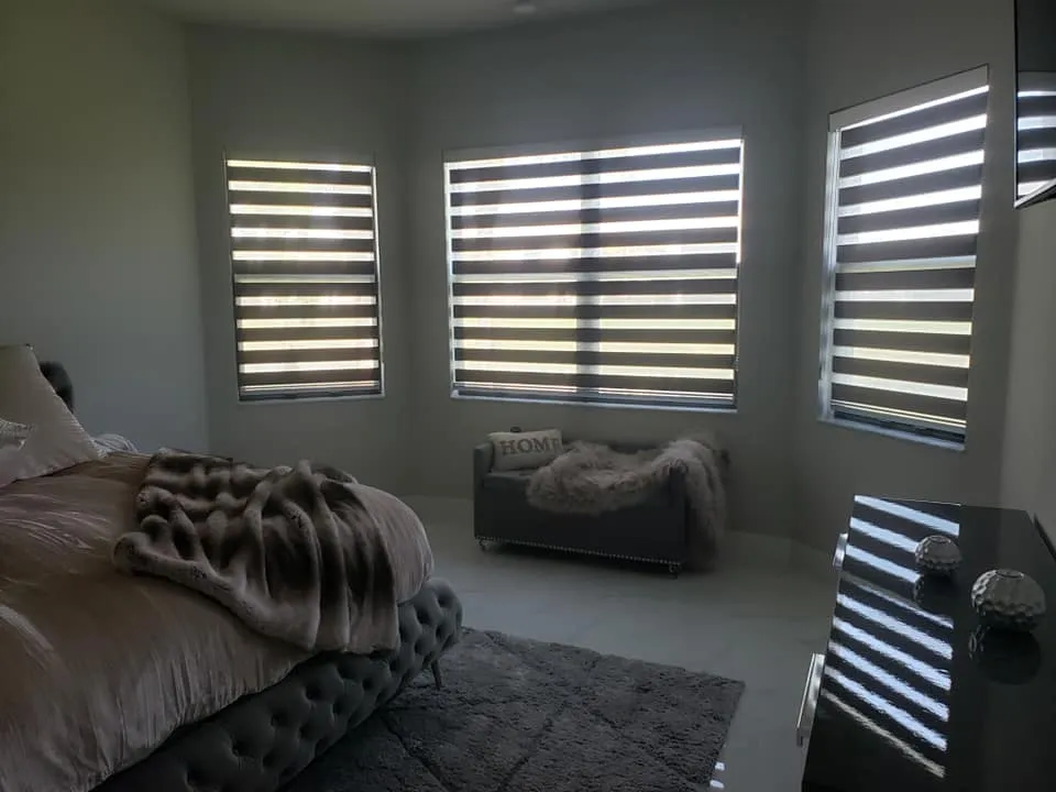 A modern bedroom with large windows covered by striped blinds, a tufted bed with a fur blanket, a small sofa with a Home pillow, and a black dresser with decorative items. Soft, natural light fills the room.