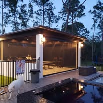 A modern outdoor covered patio with mesh screens lit by warm lights, adjacent to a pool. A white dog stands near a fence, and tall trees are visible in the background at dusk.
