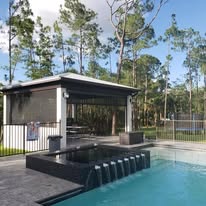 A modern outdoor pool with a small waterfall feature, surrounded by a black fence. In the background, there is a covered patio area and tall pine trees under a partly cloudy sky.