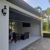 Covered patio with outdoor seating, a mounted TV, and a wall lantern on a white house exterior. Trees and a fence are visible in the background under a blue sky.