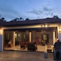 Patio view of a house at dusk, with a large glass sliding door revealing a well-lit living area with chairs and tables inside. A person sits outside on the right near a small bin. The sky is partly cloudy.