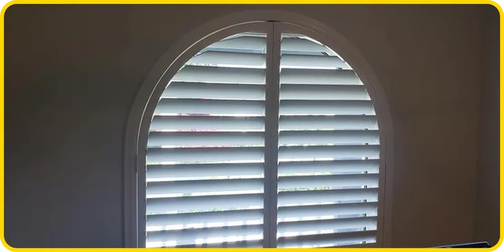 plantation shutters A white arched window with horizontal blinds partially open, allowing some light to filter into the room. The window is set in a plain wall with a yellow border around the image.