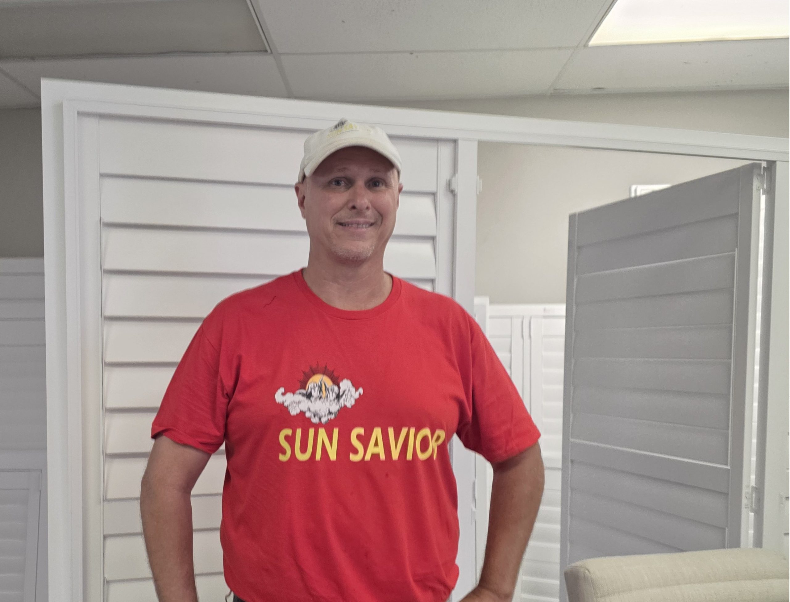 The Boval Family Chris Irminger wearing a red SUN SAVIOR T-shirt and a white cap stands indoors in front of sleek motorized shades Ft Myers, smiling at the camera.