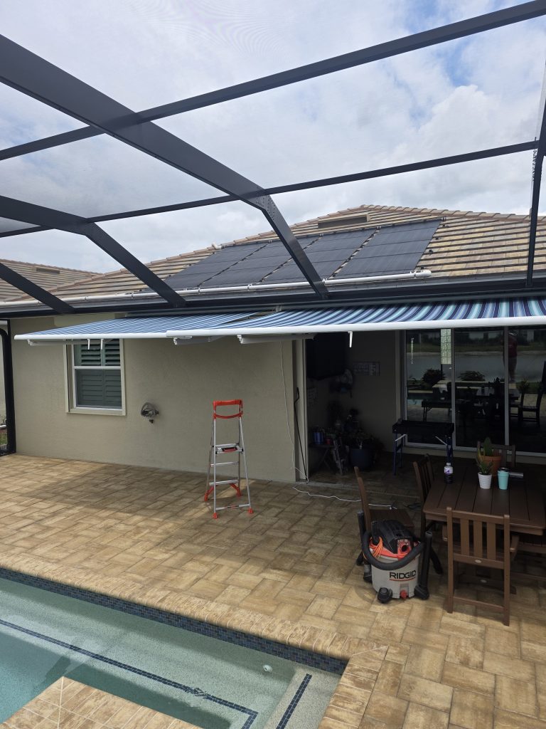 A backyard patio with a pool, paver flooring, and a screened enclosure features a Gallery Central Florida Awnings retractable striped awning. A red step ladder and shop vacuum are nearby, along with a table and chairs on the patio.