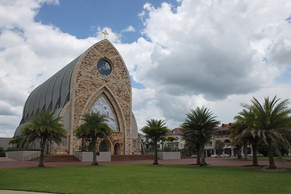 ave maria fl A modern stone church with a large arched entrance, round window, and cross on top features retractable awnings. Surrounded by palm trees and green grass under a partly cloudy sky in Bradenton, FL.