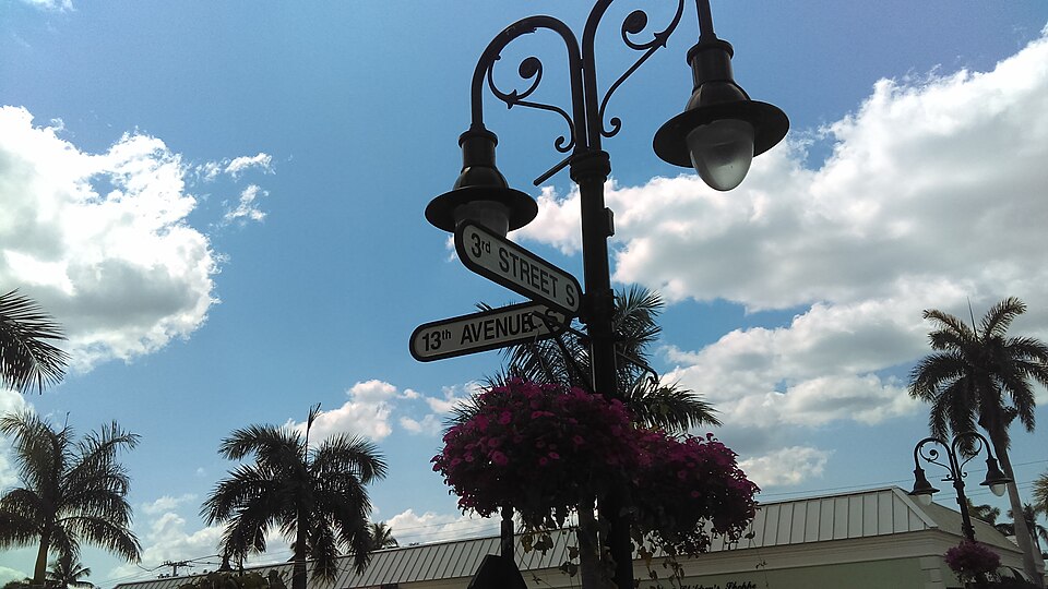 A street sign at the intersection of 3rd Street and 13th Avenue, with hanging flowers, palm trees, retractable awnings in Naples FL, and a partly cloudy sky in the background.
