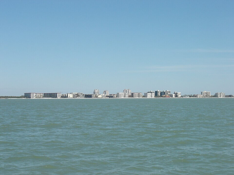 bonita springs A view of a calm, blue sea with gentle waves in the foreground and a distant shoreline lined with modern high-rise buildings, some featuring retractable awnings Bradenton FL, under a clear, blue sky.