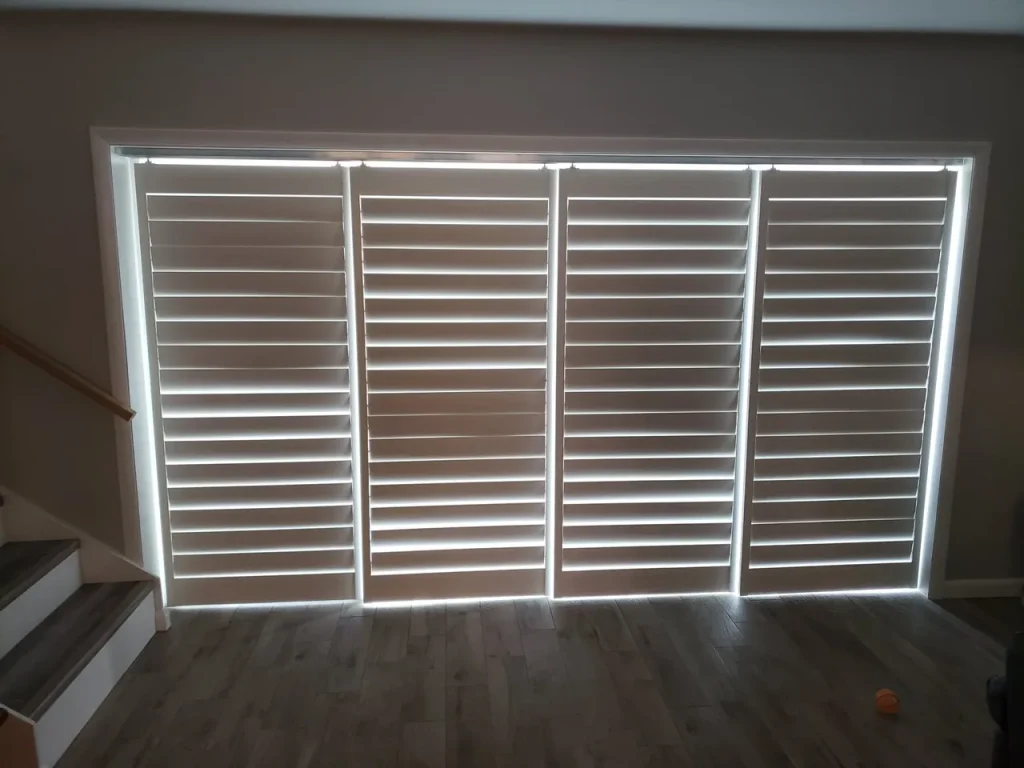 A set of four large white plantation shutters, partially closed, with sunlight shining through the gaps. Installed by Gallery Central Florida Awnings, they cover a wide window above a wooden floor.