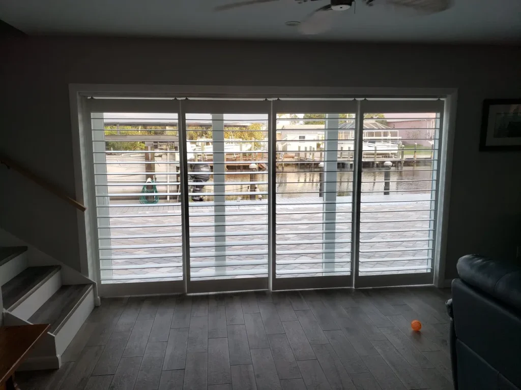 Large glass sliding doors with white frames open to a waterfront deck, featuring boats docked outside. The room has gray tile flooring, a left-side staircase, and part of a blue chair—showcasing Gallery Central Florida Awnings style.