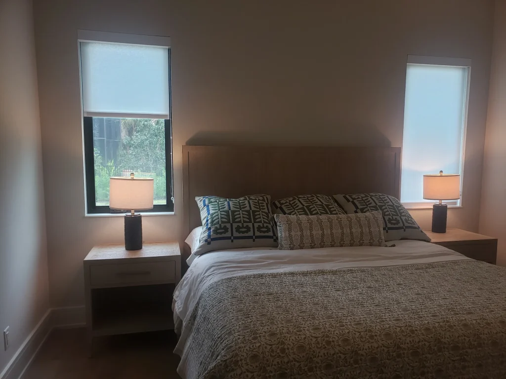 A neatly made bed with patterned pillows and a textured blanket is centered between two nightstands, each holding a lit lamp. Gallery Central Florida Awnings filter natural light through the windows with blinds on either side of the bed.