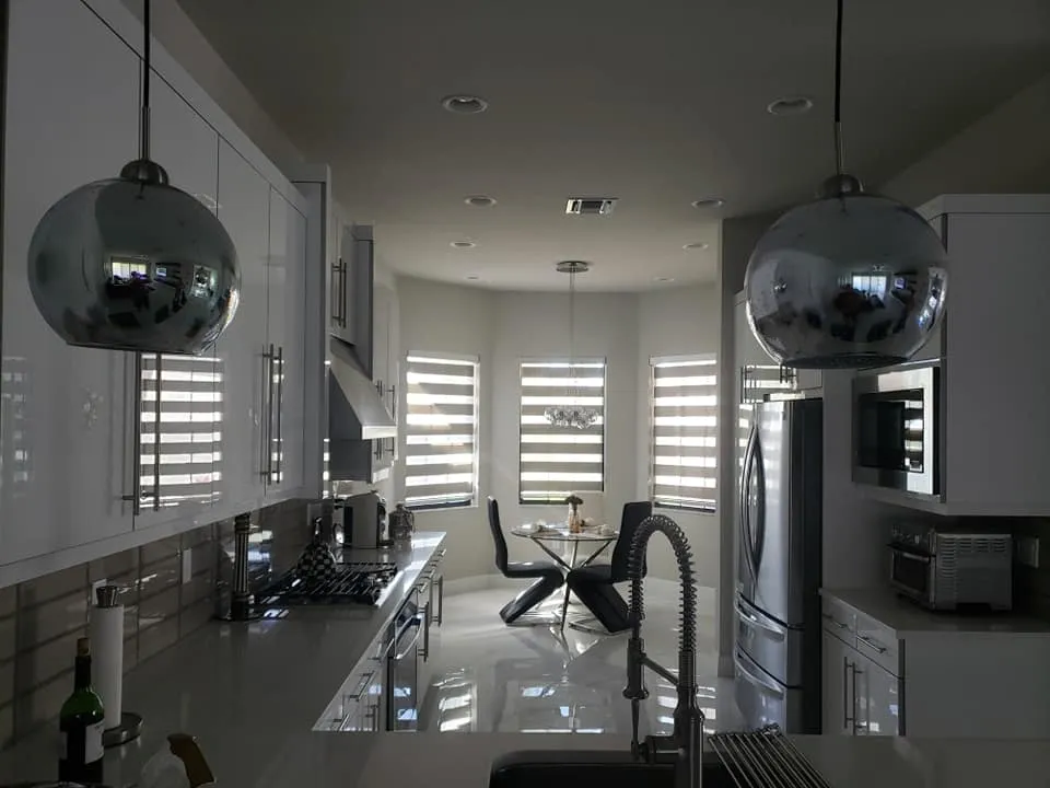 Modern kitchen with white cabinets, stainless steel appliances, and two reflective pendant lights. In the background, a dining table by large windows features striped blinds from Gallery Central Florida Awnings, letting in sunlight.