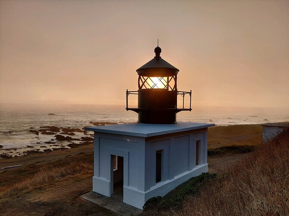 A lighthouse on a grassy cliff overlooks the ocean at sunset, its light glowing brightly as waves roll in under an orange sky—much like homes with retractable awnings in Punta Gorda FL, blending beauty and function by the coast.