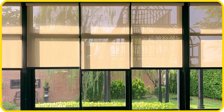 Yellow roller blinds from Gallery Central Florida Awnings partially cover large windows, revealing a garden outside with brick walls, green bushes, tall plants, and black metal structures.