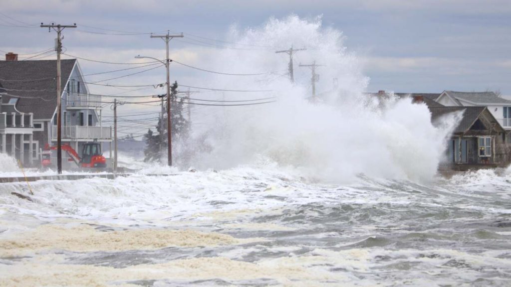Large ocean waves crash against a coastal road lined with houses and utility poles, many featuring Gallery Central Florida Awnings, as heavy spray and flooding occur under a cloudy sky.
